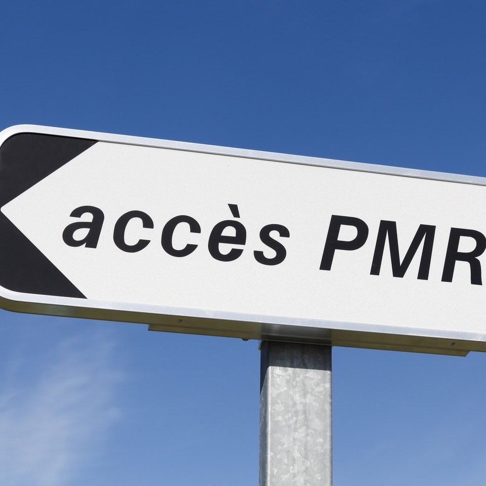 Panneau directionnel blanc indiquant 'accès PMR' (Personnes à Mobilité Réduite) avec une flèche noire vers la gauche.
