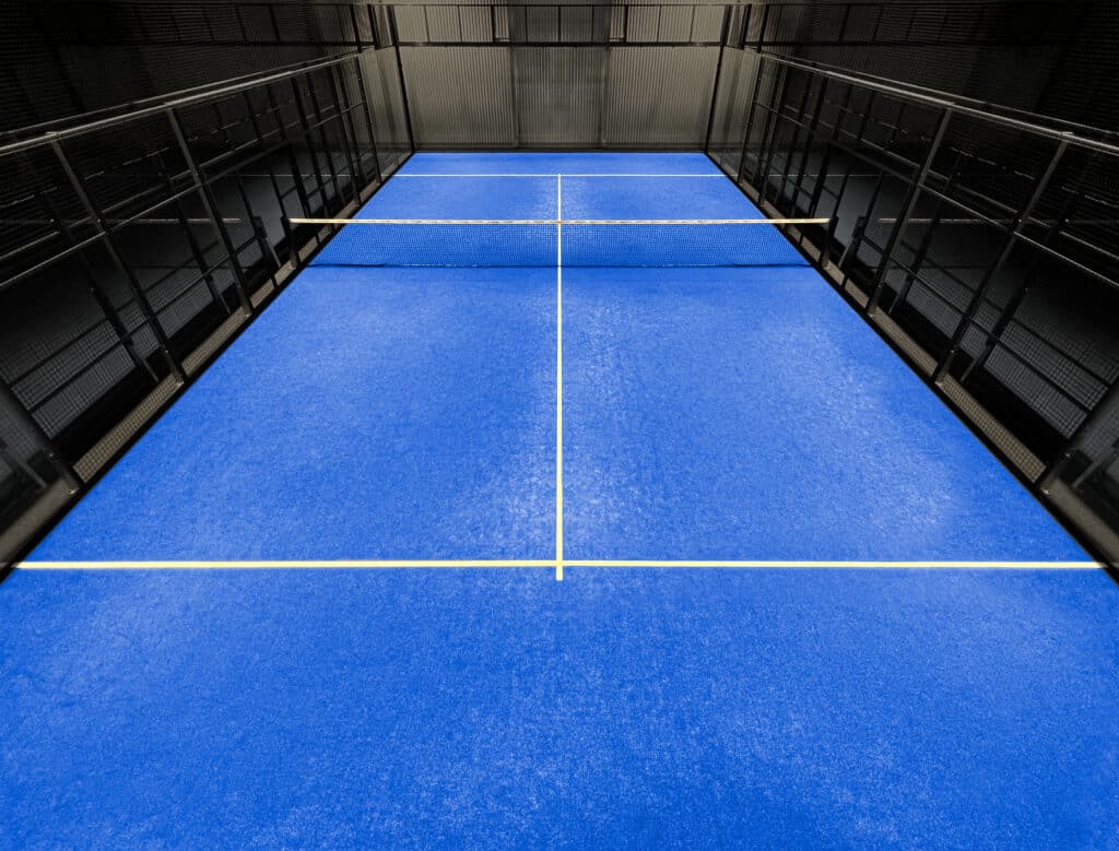 Court de padel intérieur bleu vif avec lignes blanches et filet central. Entouré de murs grillagés noirs et d'un plafond métallique.