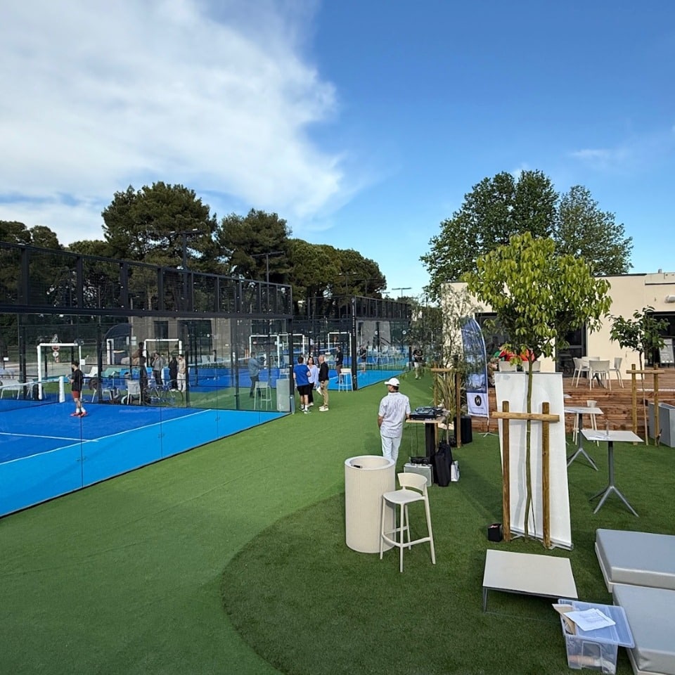 Courts de padel bleus avec joueurs, public et espace lounge extérieur sur gazon vert. Ambiance conviviale sous ciel bleu.