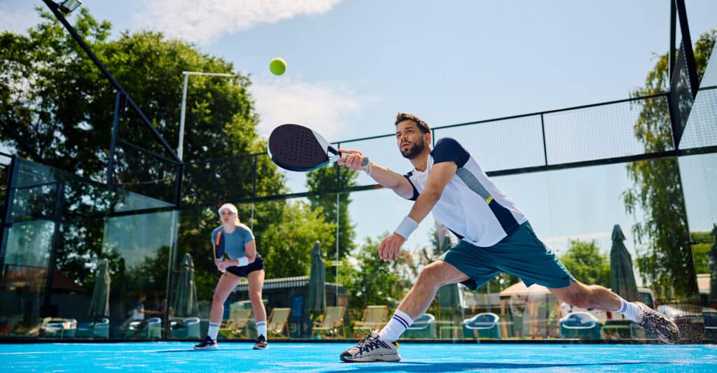 Joueur de padel masculin en pleine action, lungeant pour frapper la balle sur un court bleu. Une joueuse est en arrière-plan.