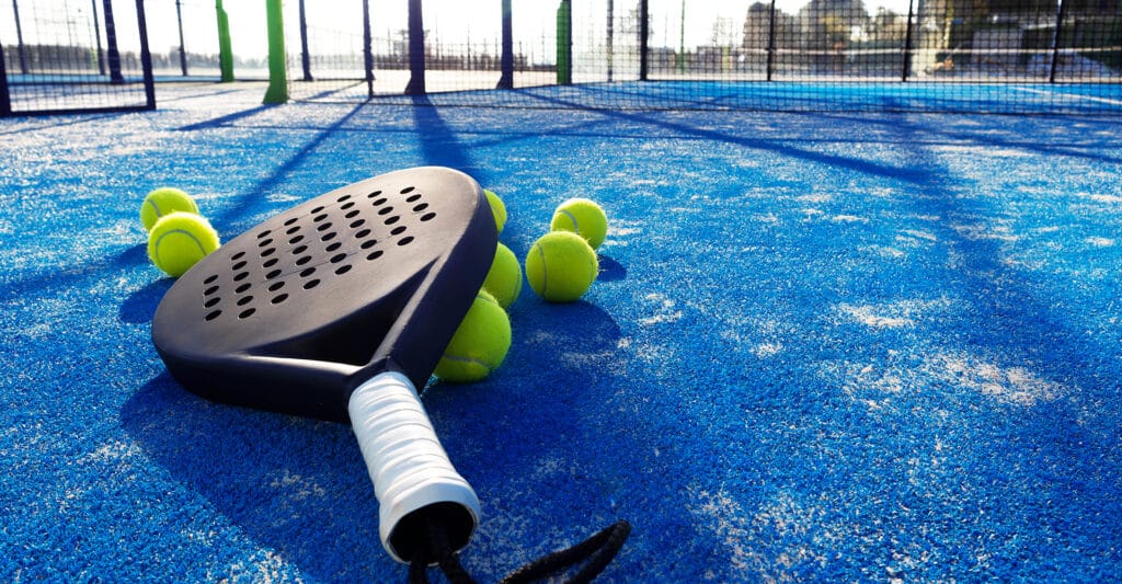 Raquette de padel noire et balles jaunes sur un terrain bleu au soleil.